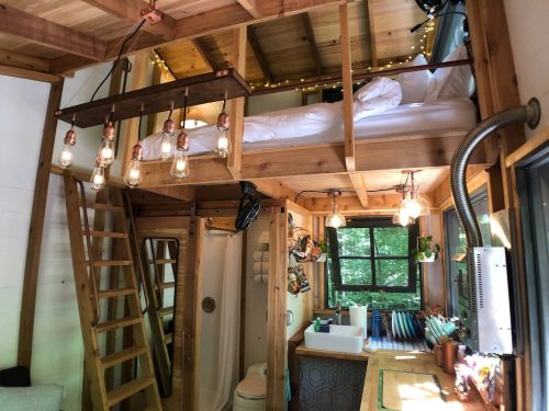 Cozy tiny home interior featuring a lofted bed, wooden accents, and a compact kitchen with a ladder and hanging lights.