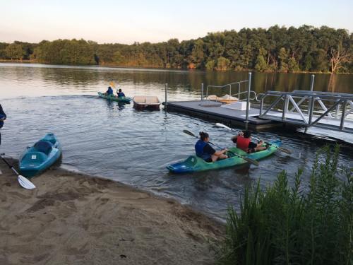 Two kayakers paddle on a calm lake near a sandy shore and a dock, surrounded by trees in the background.