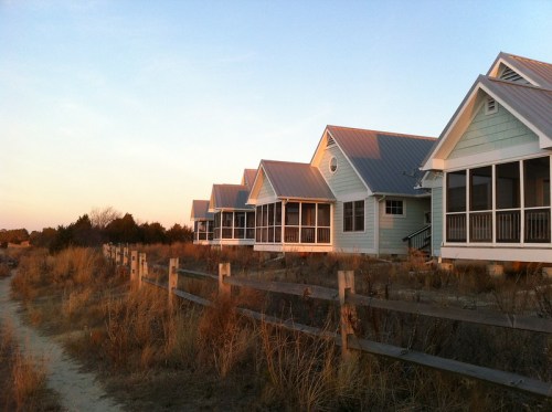 Row of beach houses with porches, set against a sunset sky and surrounded by tall grass and a wooden fence.
