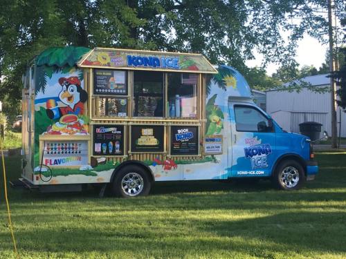 A colorful Kona Ice truck parked on grass, featuring tropical designs and a menu of flavored shaved ice treats.
