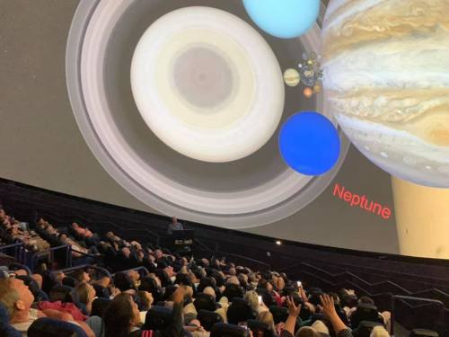Audience seated in a planetarium, viewing a projection of planets, including Jupiter and Neptune, on a dome screen.
