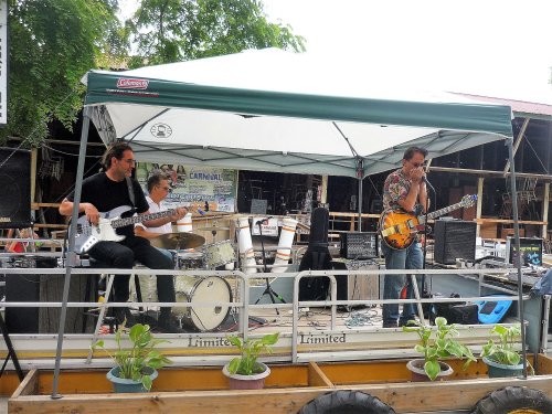 A band performs on a stage under a canopy, featuring a guitarist, bassist, and drummer, with plants in the foreground.