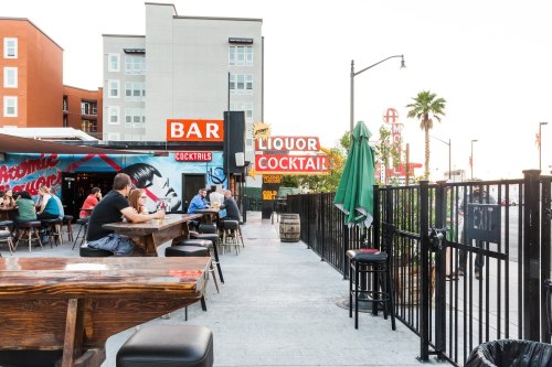 Outdoor bar scene with people seated at tables, colorful signs, and palm trees in the background.