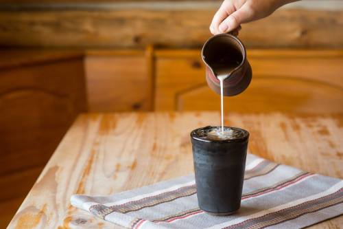 A hand pours milk from a small pitcher into a black cup on a wooden table with a striped cloth.