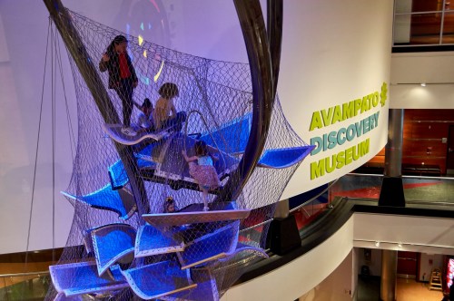 Children play on a colorful net structure inside the Avampato Discovery Museum, with vibrant walls in the background.