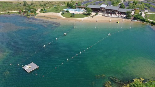 Aerial view of a lake with a sandy beach, swimming area, and a building with a pool in the background.