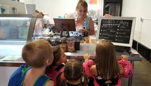 A woman serves ice cream to excited children at a shop, with various toppings displayed in jars.