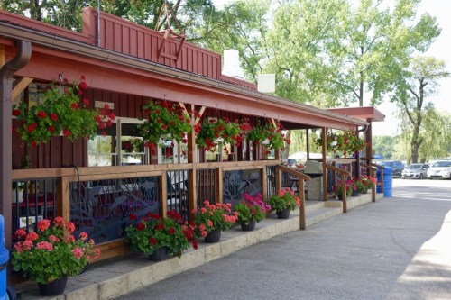 A charming restaurant exterior with vibrant flower baskets and planters, surrounded by trees and parked cars.
