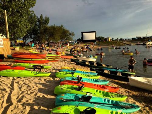 Colorful kayaks lined up on a sandy beach, with people enjoying the water and a large screen set up for a movie.