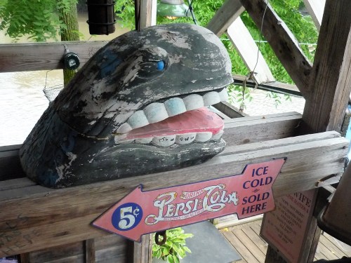 A weathered whale sculpture with a painted mouth, next to a sign advertising ice-cold soda for five cents.