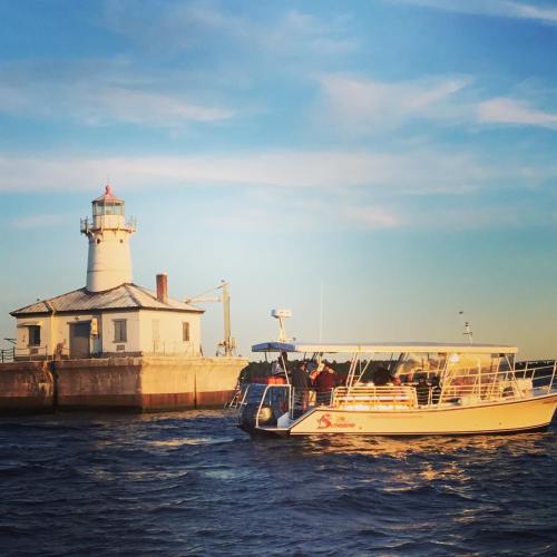 A boat passes by a lighthouse on a calm waterway during sunset, with a clear blue sky in the background.