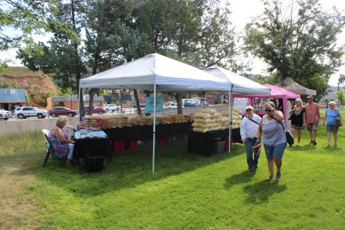 A market scene with tents selling goods, people walking by, and greenery in the background.