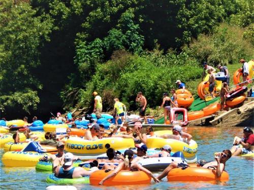 A lively scene of people relaxing on colorful inner tubes in a river, surrounded by greenery and sunny weather.
