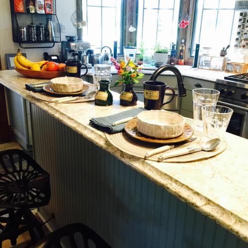 A cozy kitchen scene with a marble countertop, two place settings, fresh fruit, and a small flower arrangement.