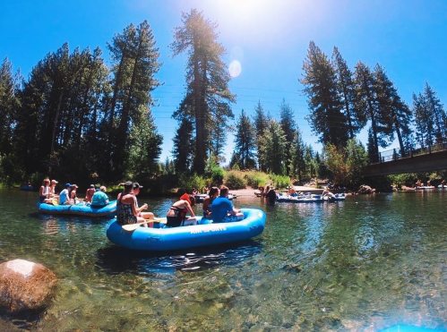 People enjoying a sunny day floating on blue rafts in a clear river surrounded by tall trees.