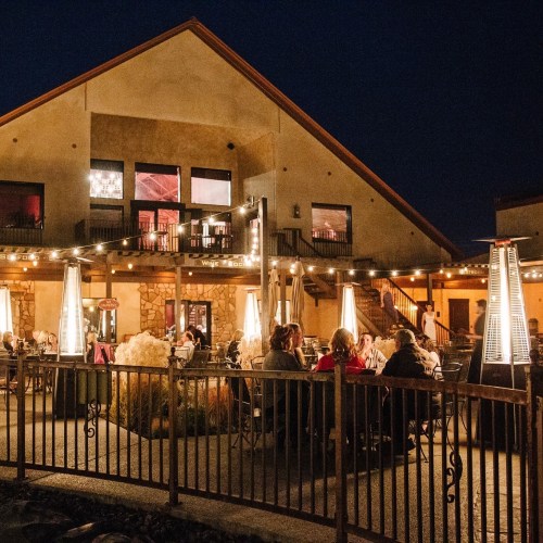 A cozy restaurant patio at night, filled with diners and warm lights, surrounded by a rustic building.