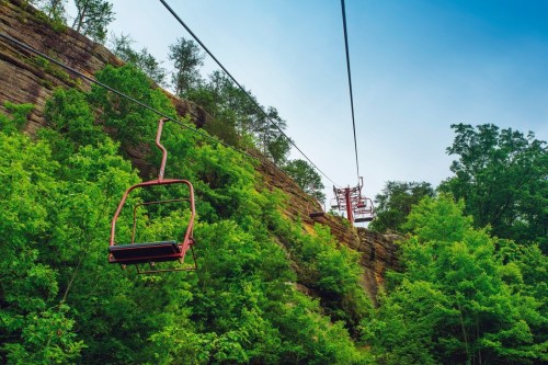 A red chairlift ascends a rocky hillside, surrounded by lush green trees under a clear blue sky.