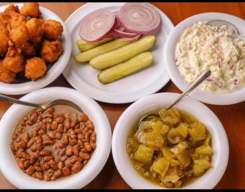 A spread of food including fried snacks, pickles, onions, coleslaw, beans, and a vegetable dish on white plates.