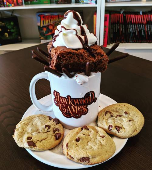 A mug topped with a brownie, whipped cream, and chocolate drizzle, surrounded by chocolate chip cookies on a plate.