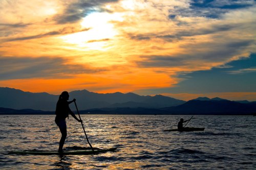 Silhouettes of two people paddleboarding at sunset, with mountains and colorful clouds in the background.
