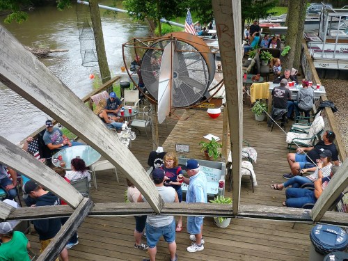 A lively riverside gathering with people socializing, sitting on a deck, and enjoying drinks under trees.