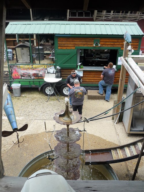 A rustic outdoor food stand with a fountain in the foreground and two people interacting near the food truck.