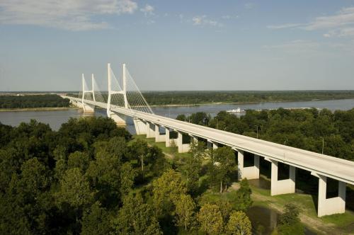 A modern cable-stayed bridge spans a river, surrounded by lush greenery and a clear blue sky.