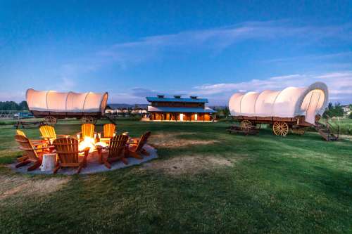 A cozy outdoor scene featuring a fire pit surrounded by wooden chairs, with two covered wagons and a lodge in the background.