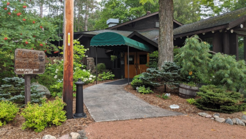 A welcoming entrance to a rustic building surrounded by greenery and decorative plants, with a sign indicating hours.