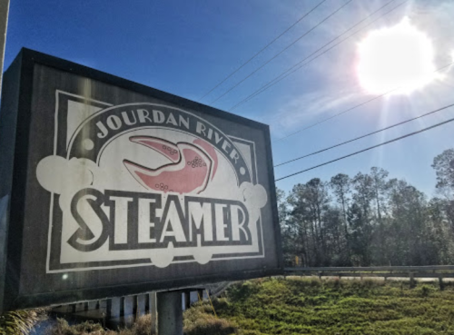 Sign for Jourdan River Steamer featuring a crab graphic, with a bright sun and trees in the background.