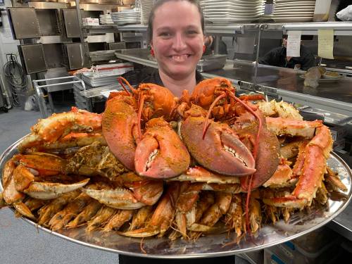 A smiling chef holds a large platter of various cooked seafood, including lobsters and shrimp, in a kitchen setting.