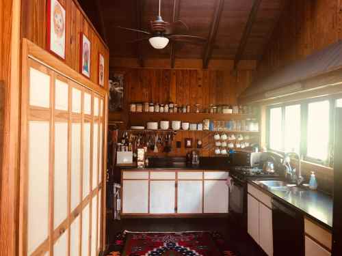 Cozy wooden kitchen with shelves of dishes, a stove, and a colorful rug on the floor. Bright natural light from windows.