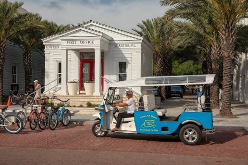 A blue electric vehicle parked near a white post office building, with bicycles nearby and palm trees in the background.