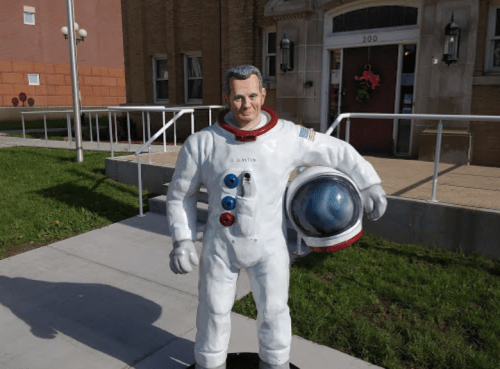 A life-sized astronaut statue holding a helmet, standing on a sidewalk in front of a building with flowers.