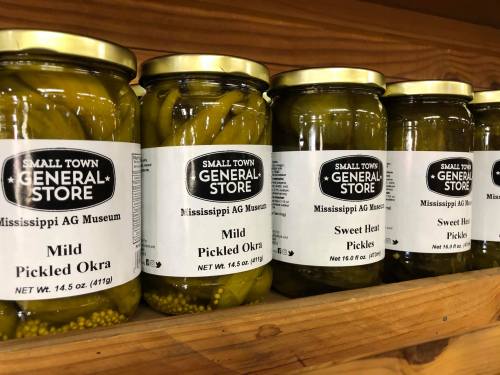 Jars of pickled okra and pickles on a wooden shelf, labeled with "Small Town General Store" and "Mississippi AG Museum."