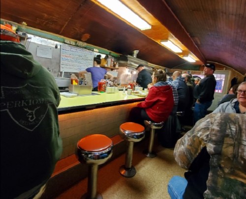 A cozy diner scene with patrons seated at a counter, chatting and enjoying food, while staff prepare meals in the background.