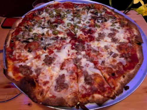 A large pizza with various toppings, including sausage, olives, and mushrooms, sliced into equal pieces on a metal tray.