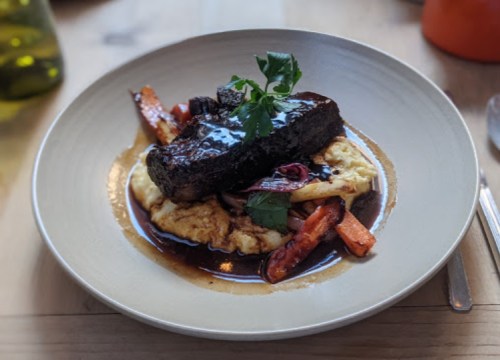 A plate of braised beef on creamy polenta, garnished with herbs and accompanied by roasted carrots and onions.