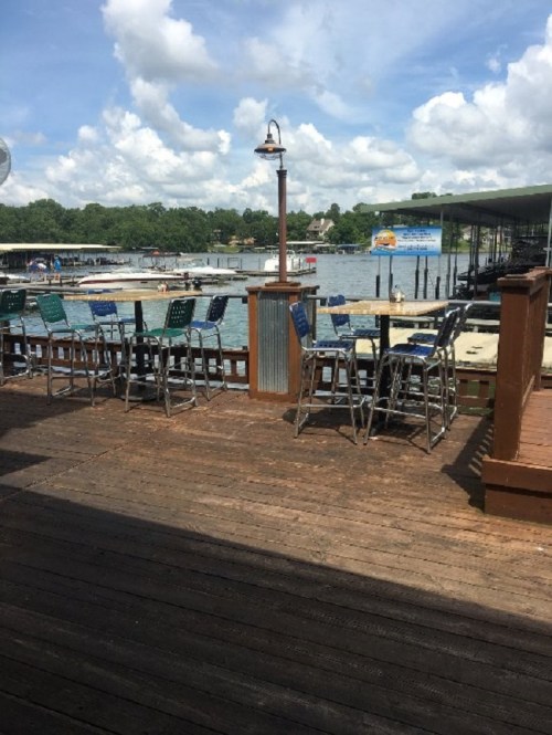 View of a lakeside deck with empty bar stools, boats in the water, and a cloudy sky above.