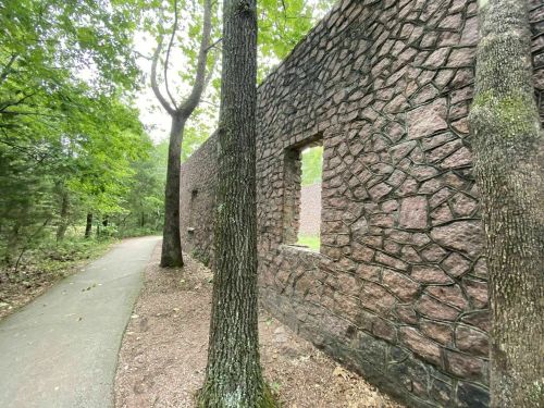 A stone wall with a window opening, surrounded by trees, next to a paved path in a wooded area.