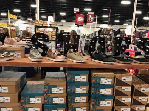 A display of various shoes on a wooden table, with a large "SHOES" sign in the background at a market.