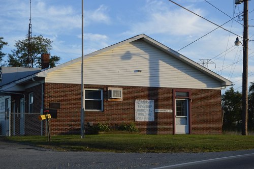 A brick municipal building with a sign, mailbox, and grassy area, set against a clear sky.
