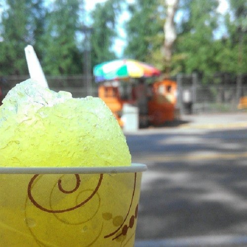 A close-up of a yellow snow cone in a cup, with a colorful vendor cart in the background.