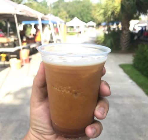 A hand holding a clear cup of iced coffee, with a market scene in the background.