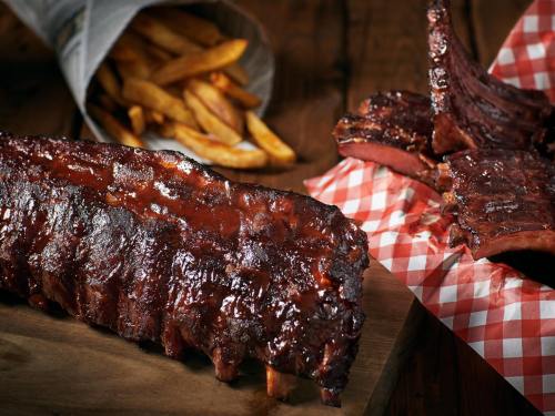 A close-up of glazed ribs on a wooden board, with a side of fries in a paper cone.