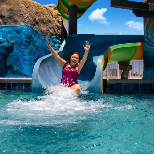A girl in a swimsuit joyfully slides down a water slide into a pool, splashing water around her.