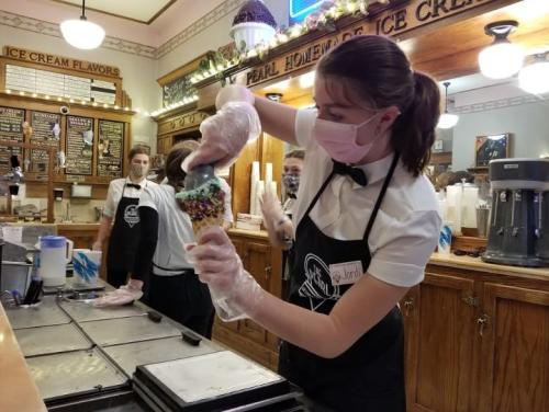 A server in a mask and gloves prepares an ice cream cone at a vintage ice cream shop, with staff in the background.