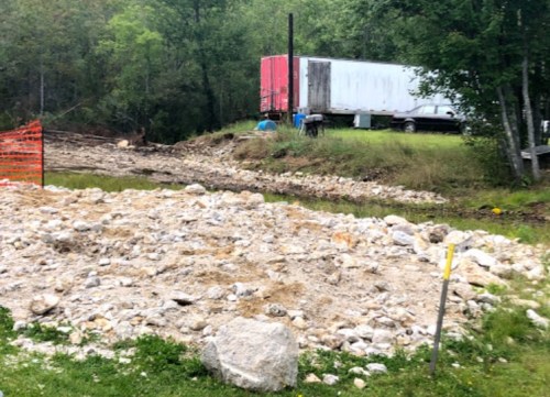 A gravelly area with a pile of rocks, a trailer, and a car in a wooded setting, with orange construction fencing nearby.