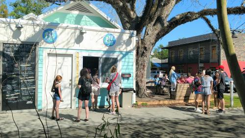 A colorful food stand with people waiting in line, surrounded by trees and buildings on a sunny day.