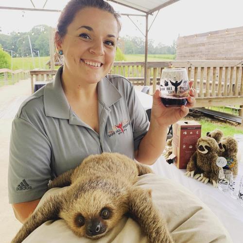 A smiling woman holds a drink while a sloth rests on her lap at an outdoor event.